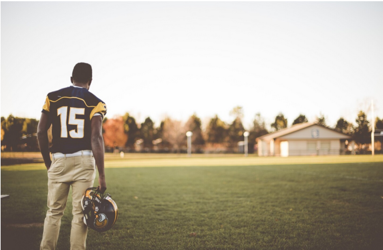 Football player holding helmet, looking out over a grassy field